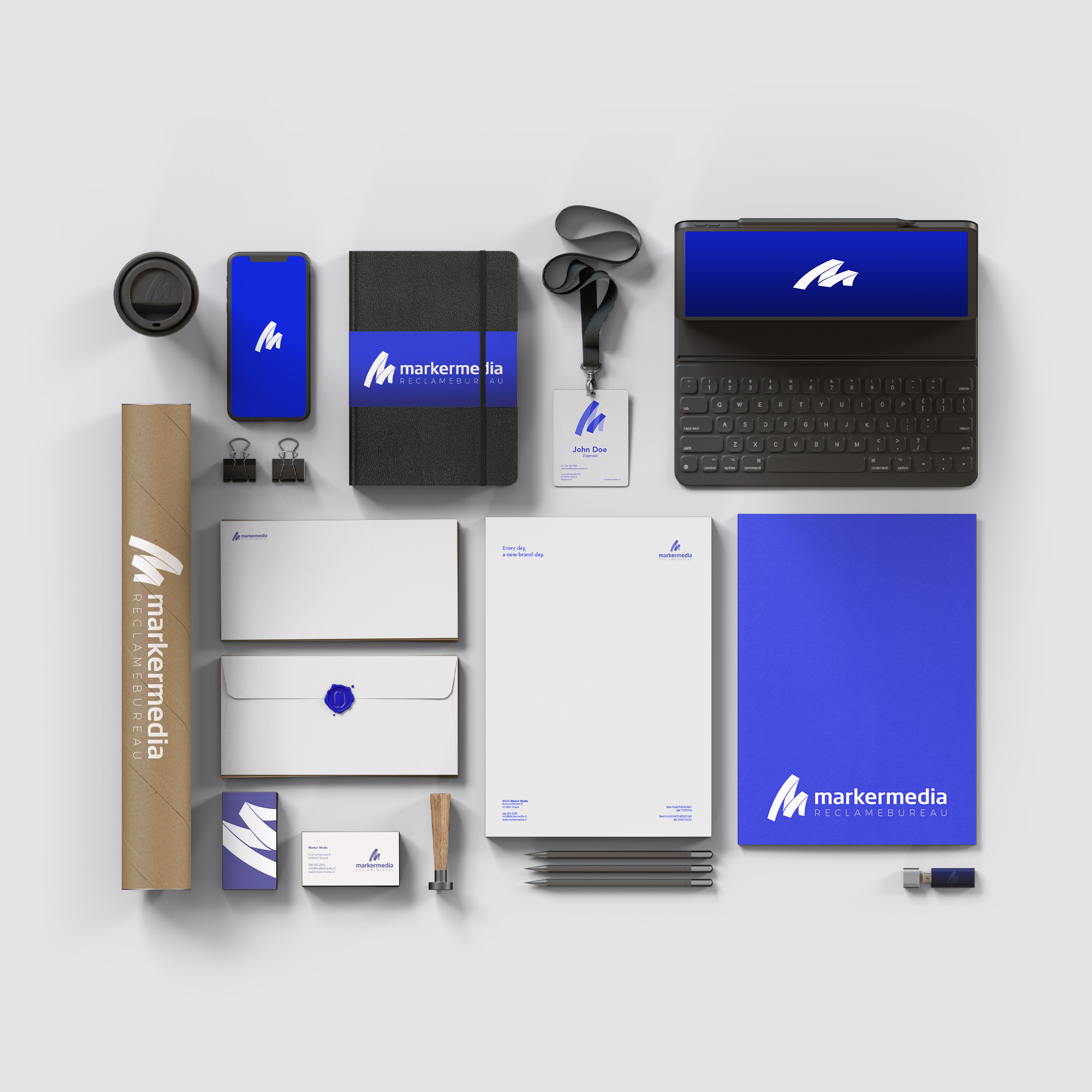 Een overzicht van de huisstijl van Marker Media, met verschillende merkitems zoals notitieboekjes, pennen, visitekaartjes, enveloppen, en een smartphone met het logo van Marker Media, allemaal gepresenteerd op een vlakke ondergrond voor een professionele uitstraling.
